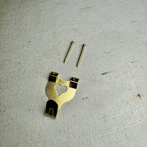 May include: A gold-colored metal picture hook with a heart-shaped cutout. Two gold-colored nails are included.