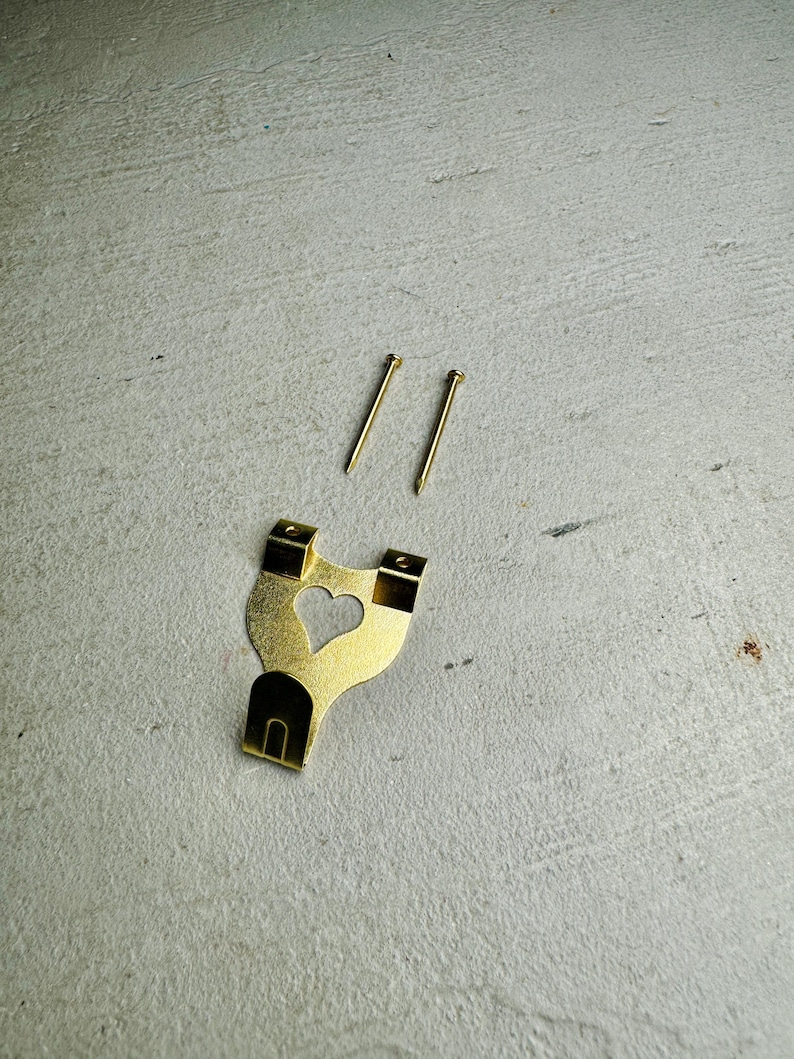 May include: A gold-colored metal picture hook with a heart-shaped cutout. Two gold-colored nails are included.