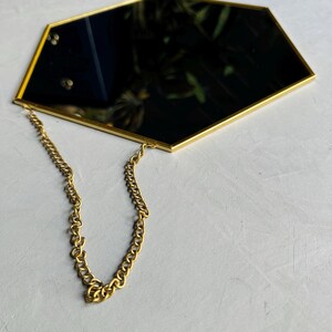 May include: A gold hexagonal mirror with a chain hanging from the top. The mirror is reflecting a green plant.