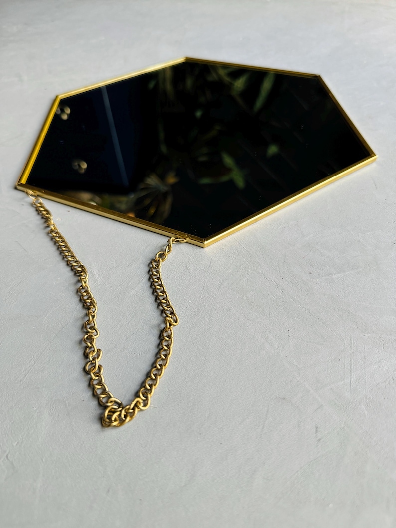May include: A gold hexagonal mirror with a chain hanging from the top. The mirror is reflecting a green plant.