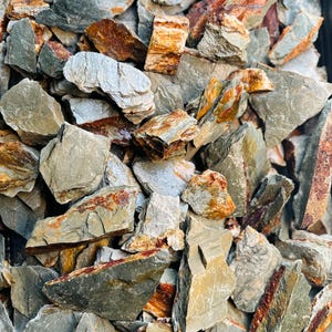 May include: A collection of irregularly shaped rocks in shades of grey, brown, and orange. The rocks have rough, textured surfaces and vary in size. Some rocks display layers of colour, creating a natural, earthy appearance. Suitable for landscaping or decorative use.