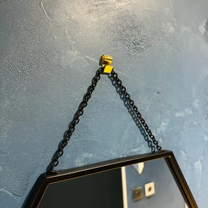 May include: A black hexagonal mirror with a black chain hanging from a gold hook on a blue textured wall.