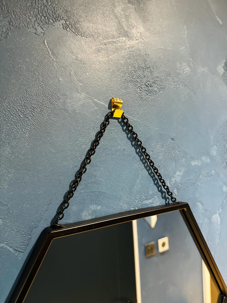 May include: A black hexagonal mirror with a black chain hanging from a gold hook on a blue textured wall.