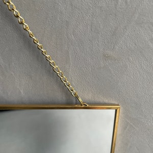May include: A gold-framed mirror with a chain hanging from the top. The mirror is rectangular and has a simple, modern design.
