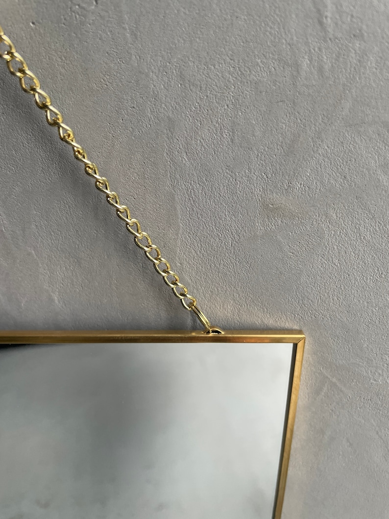 May include: A gold-framed mirror with a chain hanging from the top. The mirror is rectangular and has a simple, modern design.