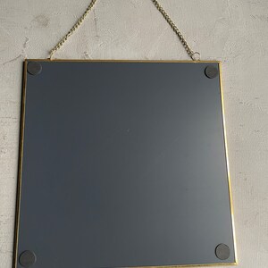 May include: A gold-framed square mirror with a dark gray backing. The mirror is hanging from a gold chain.