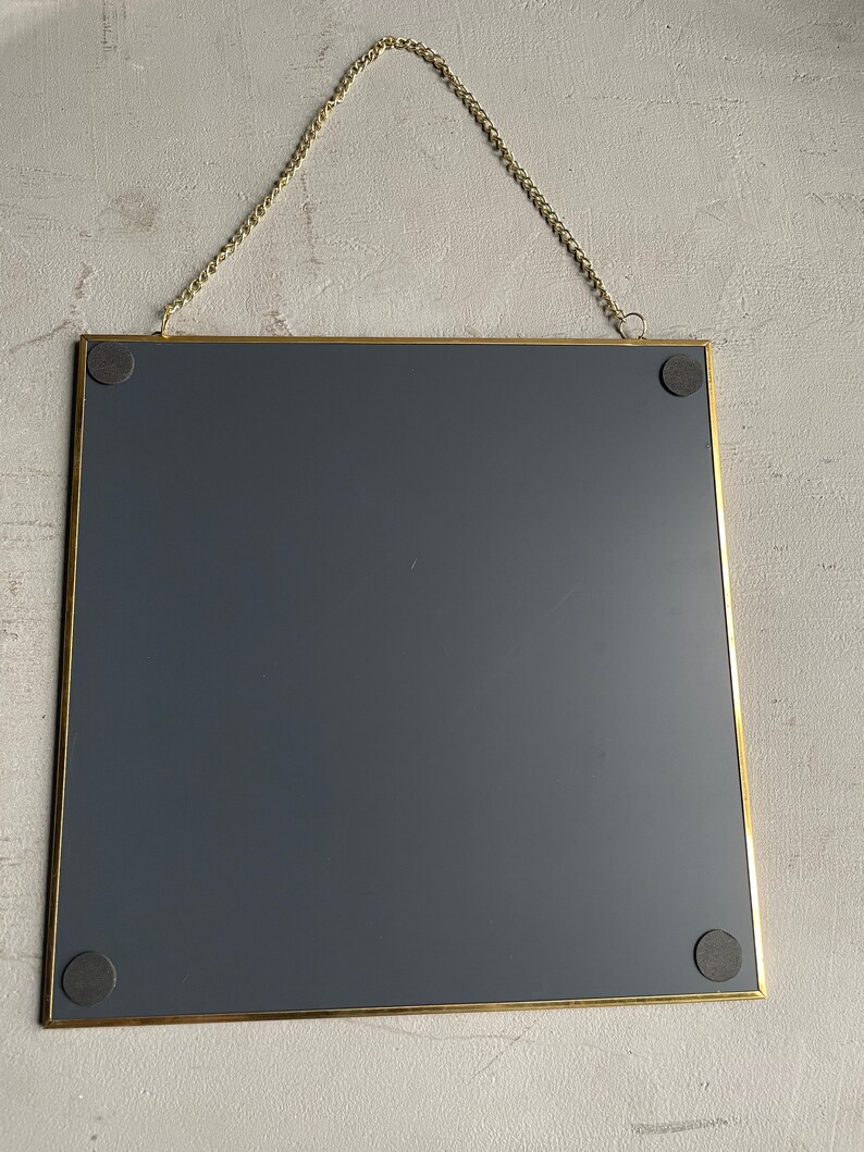 May include: A gold-framed square mirror with a dark gray backing. The mirror is hanging from a gold chain.