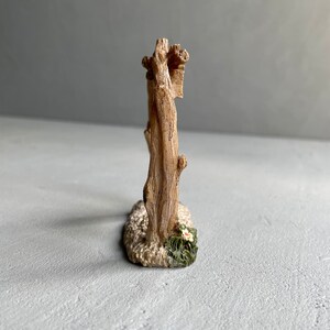 May include: A miniature wooden fence with a rustic brown finish. The fence is made of two vertical posts connected by horizontal rails. The base of the fence is decorated with white sand and green foliage with a white flower.