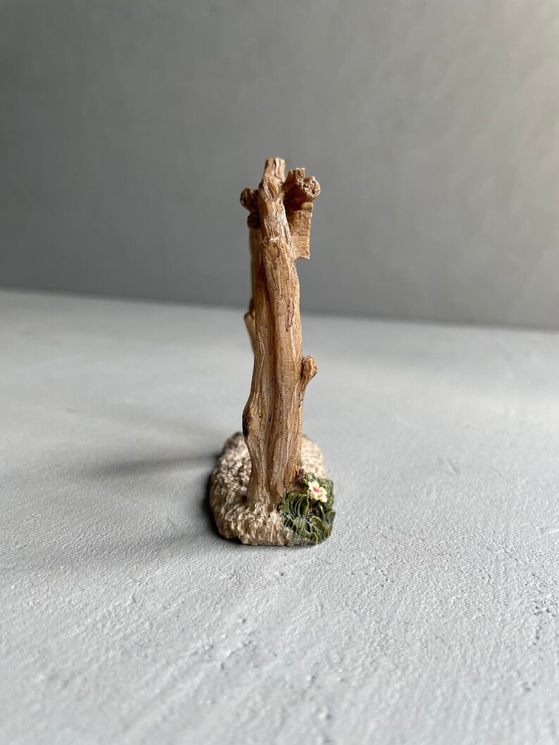 May include: A miniature wooden fence with a rustic brown finish. The fence is made of two vertical posts connected by horizontal rails. The base of the fence is decorated with white sand and green foliage with a white flower.