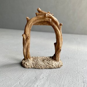 May include: A small, rustic wooden archway with a sandy base. The archway is made of intertwined branches and is perfect for adding a touch of nature to a miniature fairy garden or terrarium.