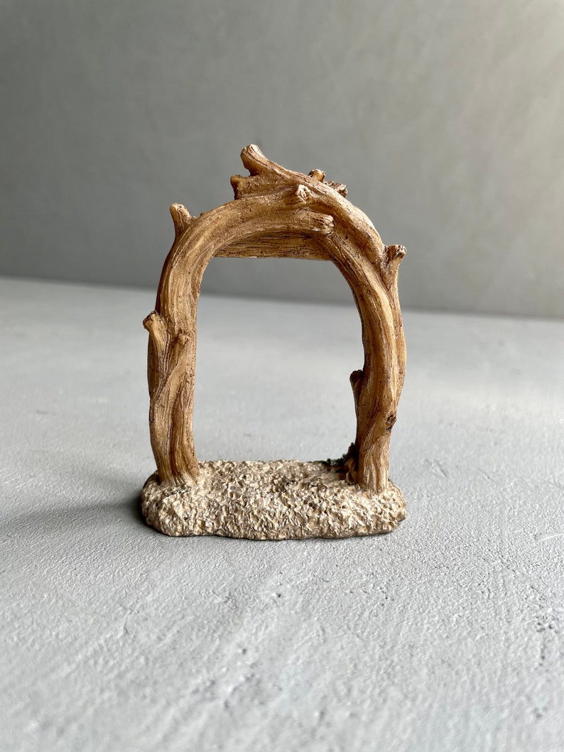 May include: A small, rustic wooden archway with a sandy base. The archway is made of intertwined branches and is perfect for adding a touch of nature to a miniature fairy garden or terrarium.