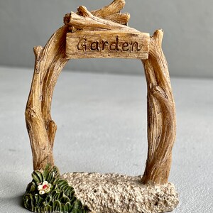 May include: A miniature wooden garden archway with a sign that reads "Garden". The archway is made of brown branches and has a small flower at the base.