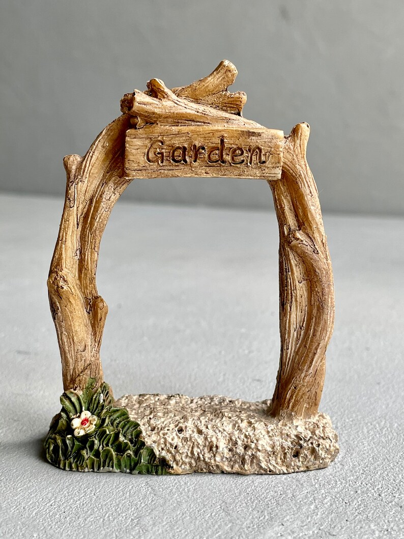 May include: A miniature wooden garden archway with a sign that reads "Garden". The archway is made of brown branches and has a small flower at the base.