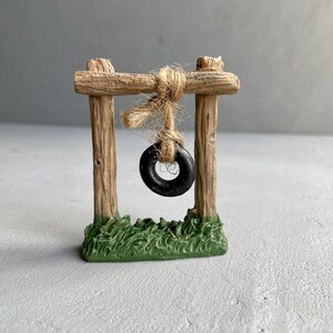 Pu&ograve; includere: Un'altalena in legno in miniatura con un pneumatico nero appeso. L'altalena &egrave; su una base di erba verde.