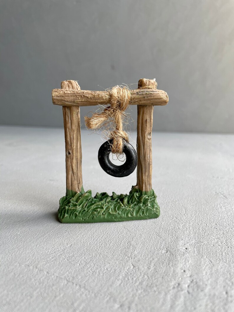 Pu&ograve; includere: Un'altalena in legno in miniatura con un pneumatico nero appeso. L'altalena &egrave; su una base di erba verde.