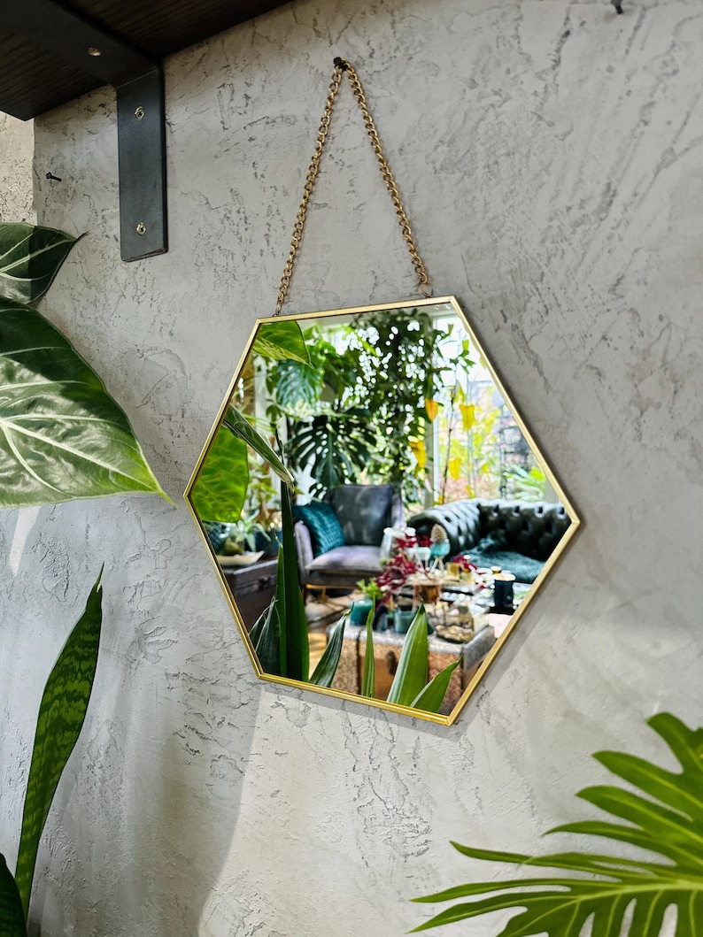 May include: A gold hexagonal mirror with a chain hanging from the top. The mirror is reflecting a room with plants and furniture.