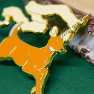 May include: An orange and gold enamel pin featuring a deer with a unicorn horn. The pin has a gold outline and accents, with a cream-colored cloud-like shape above the deer. The pin is set against a green background.