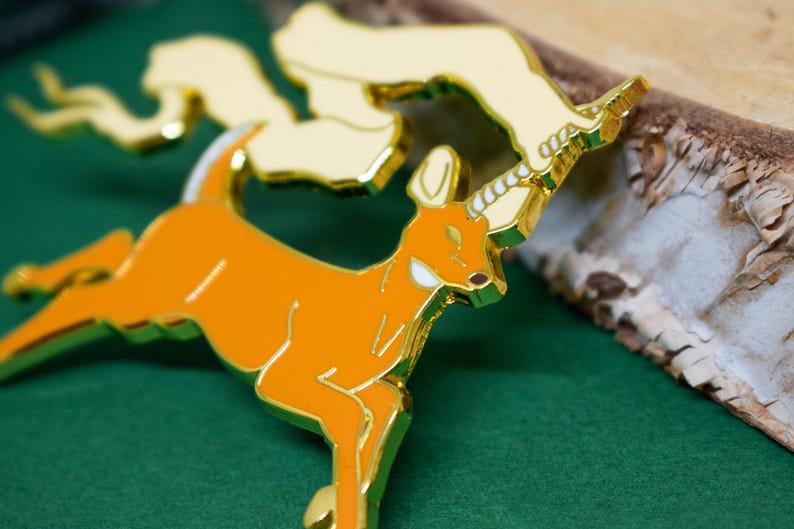 May include: An orange and gold enamel pin featuring a deer with a unicorn horn. The pin has a gold outline and accents, with a cream-colored cloud-like shape above the deer. The pin is set against a green background.