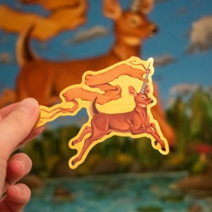 May include: A sticker of a deer with a unicorn horn, running with a yellow banner behind it. The deer is brown with a white belly and the banner is yellow with a gold trim.