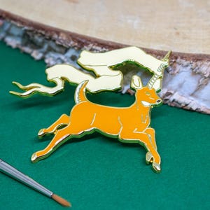 May include: An orange and gold enamel pin of a deer with a unicorn horn and a flowing banner. The pin is set against a green surface with a wooden background and a paintbrush.