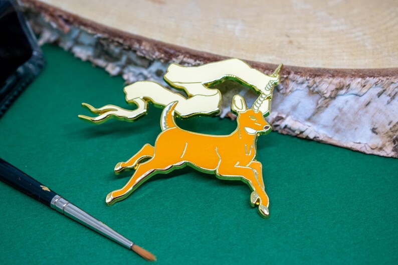 May include: An orange and gold enamel pin of a deer with a unicorn horn and a flowing banner. The pin is set against a green surface with a wooden background and a paintbrush.