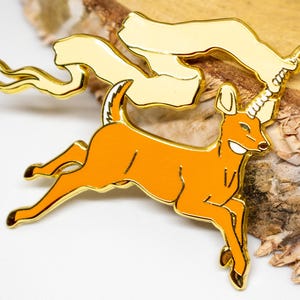 May include: A gold-colored enamel pin featuring a cartoon deer with a unicorn horn, running with a banner. The deer is orange with gold trim. The banner is cream-colored with gold trim. The pin is on a white and wood background.