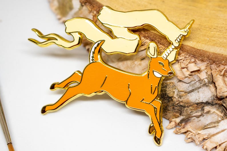 May include: A gold-colored enamel pin featuring a cartoon deer with a unicorn horn, running with a banner. The deer is orange with gold trim. The banner is cream-colored with gold trim. The pin is on a white and wood background.