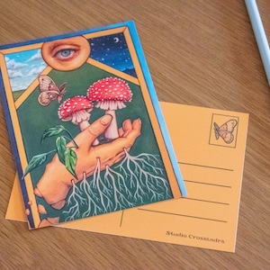 May include: A postcard featuring a hand holding three red and white mushrooms with white roots growing from the hand. The background is a green and blue landscape with a butterfly and a large eye.