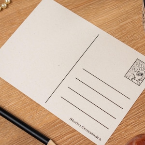 May include: A blank postcard with a black and white illustration of a hand with an eye in the palm. The postcard has lines for the address and a stamp area. The text "Studio Crossandra" is printed at the bottom.