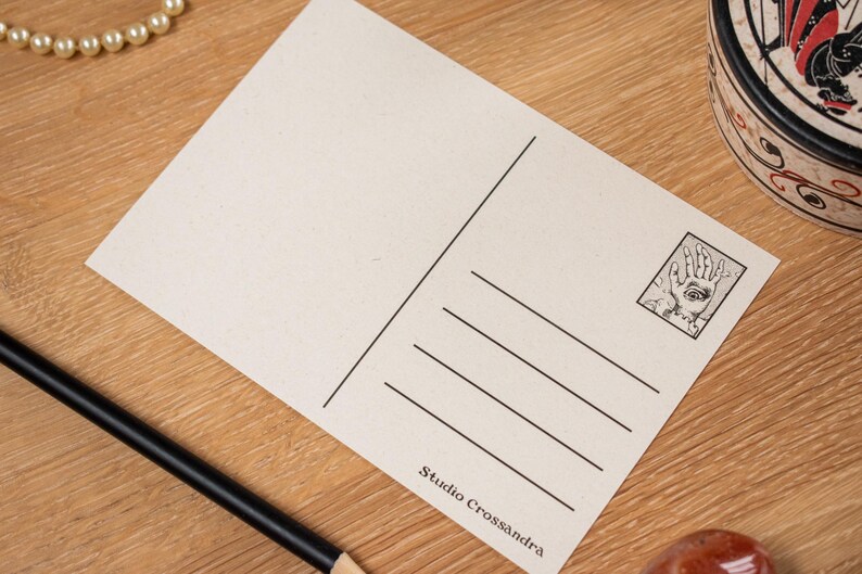 May include: A blank postcard with a black and white illustration of a hand with an eye in the palm. The postcard has lines for the address and a stamp area. The text "Studio Crossandra" is printed at the bottom.