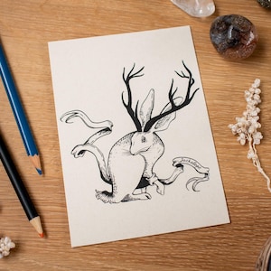 Puede incluir: Ilustración en blanco y negro de un jackalope con cuernos y una pancarta que dice "Jackalope". La obra de arte está sobre una tarjeta color crema, con lápices y objetos decorativos sobre una superficie de madera.