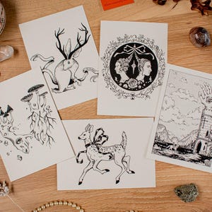 May include: A collection of black and white art prints featuring whimsical illustrations. The prints depict a deer with a bow, a rabbit with antlers, mushrooms, a tower with a hand, and portraits. The art is displayed on a wooden surface with decorative elements.