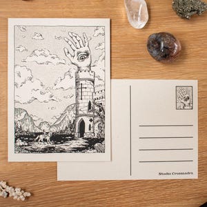 May include: Two postcards with a black and white illustration of a tower with a hand and eye on top. The front of the card shows a landscape with mountains and clouds. The back of the card is blank with lines for writing. The Studio Crossandr&eacute;'s logo is printed on the back.