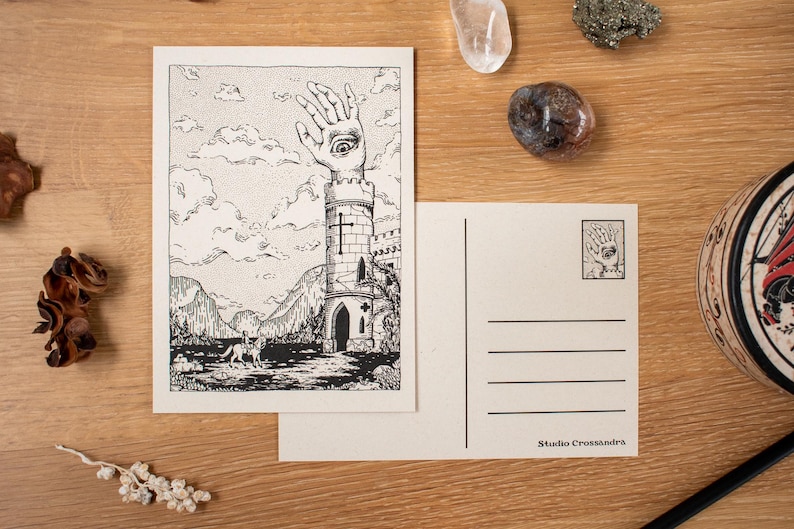 May include: Two postcards with a black and white illustration of a tower with a hand and eye on top. The front of the card shows a landscape with mountains and clouds. The back of the card is blank with lines for writing. The Studio Crossandr&eacute;'s logo is printed on the back.