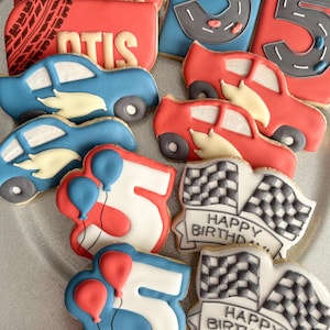 May include: A selection of decorated sugar cookies with a racing theme. Cookies feature race cars, the number 5, chequered flags, and the words "Happy Birthday" and "OTIS". The cookies are red, blue, white, and black.