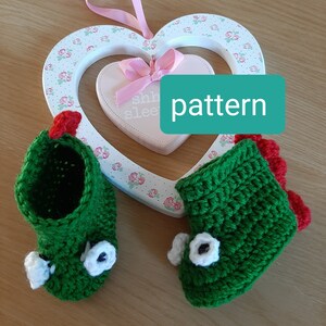 Può includere: Un paio di scarpine per neonati in crochet verde con occhi bianchi e dettagli rossi. Le scarpine sono esposte su un cartello di legno bianco a forma di cuore con il testo "Shh...slee...pattern".