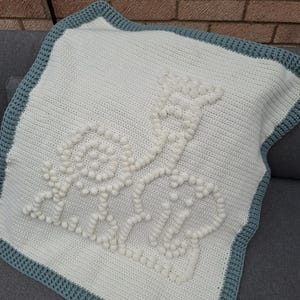 Könnte beinhalten: Eine weiße gehäkelte Babydecke mit blauem Rand. Die Decke hat ein erhabenes, strukturiertes Design einer Giraffe. Die Giraffe besteht aus weißen Knötchen, die der Decke einen 3D-Effekt verleihen.