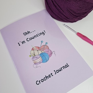 May include: A lavender Crochet Journal with the text "Shh... I'm Counting!" and "Crochet Journal". The cover features a colourful illustration of yarn balls and needles. A ball of purple yarn and a crochet hook are also visible.