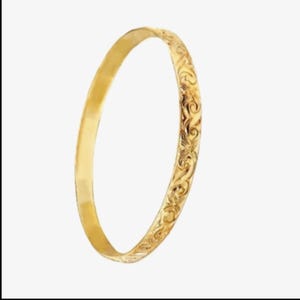 Könnte beinhalten: Ein goldfarbenes Armreif-Armband. Das Armband hat auf einer Seite eine glatte, polierte Oberfläche und auf der anderen Seite ein verziertes, eingraviertes Blumenmuster. Der Armreif ist ein vollständiger Kreis und wird vor einem weißen Hintergrund gezeigt.