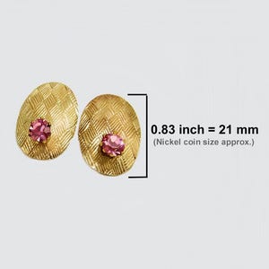 May include: A pair of gold-colored oval earrings with a textured pattern and a pink gemstone in the center. The earrings are 0.83 inches (21 mm) in length, approximately the size of a nickel.