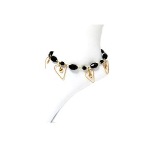 Op de afbeelding: Enkelband met zwarte facetkralen en goudkleurige hartbedels. Het enkelbandje heeft een fijne ketting en wordt gepresenteerd op een wit oppervlak. De harten zijn goud omrand en hebben een kleiner gouden hart in het midden.