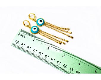 May include: A pair of gold-colored dangle earrings. Each earring features a turquoise blue circular charm with a white and black eye design. Gold chains hang from the charm. A ruler is in the image.