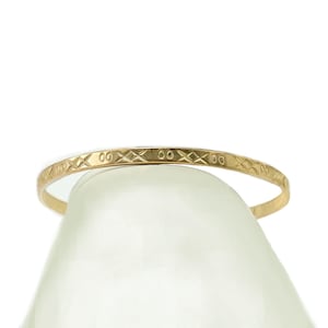 May include: A gold-colored bangle bracelet with a repeating pattern of diamond and oval shapes. The bracelet is thin and appears to be made of metal. The design is etched into the surface, creating a textured appearance. The bracelet is displayed against a white background.