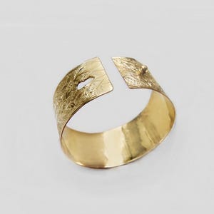 May include: A gold-toned metal bracelet with a textured surface and a small opening on one side. The bracelet is open on one side and has a small, round, gold-toned metal piece on the other side.