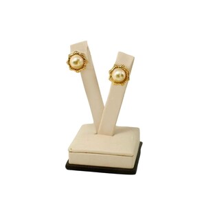 Puede incluir: Pendientes de botón dorados con una perla central y un marco decorativo en forma de sol. Los pendientes se exhiben en un expositor de joyería color crema con una base marrón oscuro, sobre un fondo blanco.