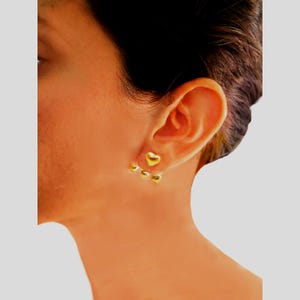 May include: A close-up of a person's ear adorned with gold heart-shaped earrings. The earrings feature a cluster of three small hearts below a larger heart, creating a cascading effect. The jewelry is a bright, shiny gold color.