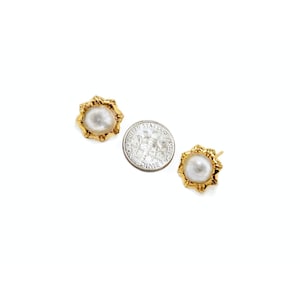 Puede incluir: Dos pendientes de botón dorados con un centro de perla y un marco texturizado en forma de sol. Una moneda de diez centavos estadounidense está al lado de los pendientes para comparar el tamaño. Los pendientes están sobre un fondo blanco.