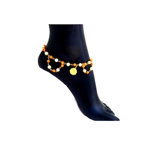 May include: Anklet with orange and white beads and a gold-coloured charm. The anklet is worn on a black foot. The beads are strung on a gold-coloured chain. The charm is a small, round pendant.