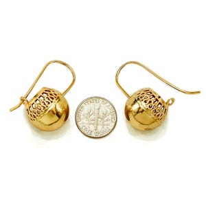 May include: A pair of gold-colored earrings with a ball-shaped design. Each earring features a hook and a textured, openwork pattern on the upper portion of the ball. A U.S. dime is placed between the earrings for size comparison.