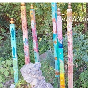 Puede incluir: Un conjunto de seis estacas de jardín de madera, altas, delgadas y de perfil cuadrado, pintadas con distintos colores y patrones, incluyendo motivos de peces, diseños florales y un rostro. Cada estaca tiene un remate redondo dorado.